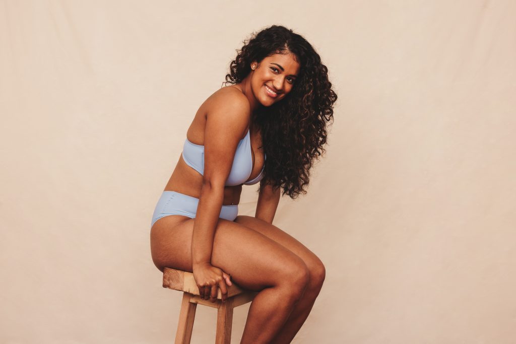 Cheerful young woman sitting comfortably in blue underwear. Happy young woman smiling at the camera while sitting alone against a studio background. Body positive young woman embracing her natural body.